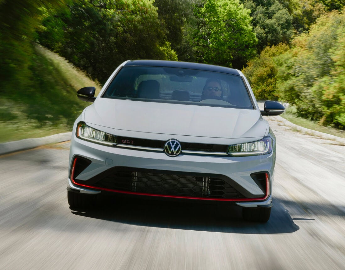 2026 Volkswagen Jetta GLI Autobahn in Monument Gray, front view driving on a winding tree-lined road.