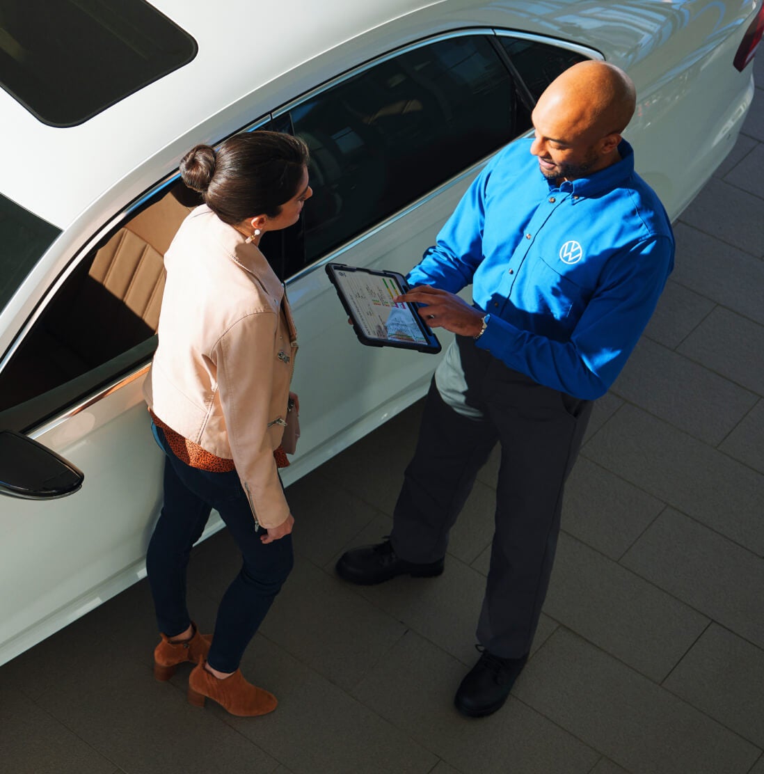 Volkswagen dealership representative shows a customer information on a tablet beside a white car, viewed from above.