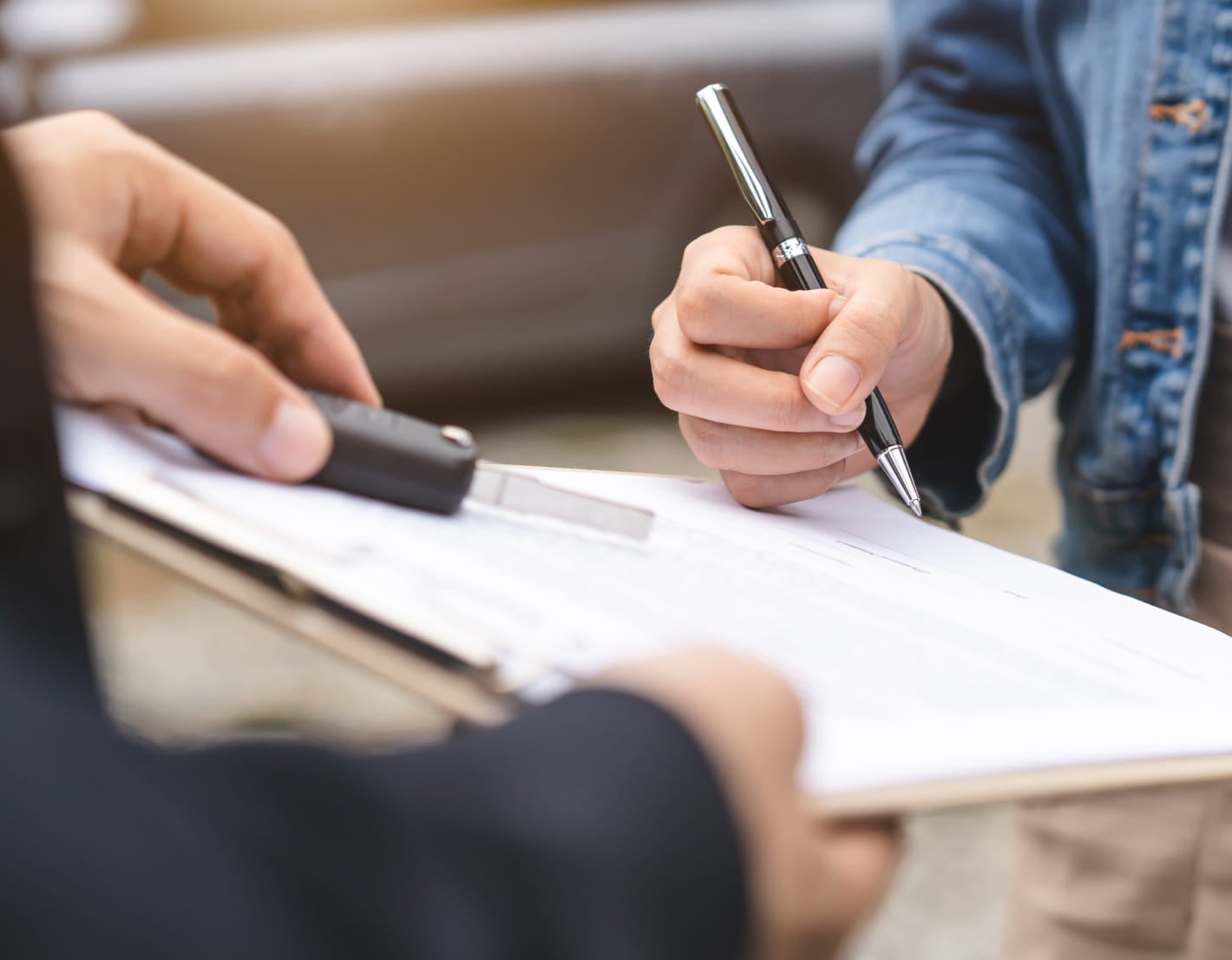 A person in denim signs a document on a clipboard, holding a pen. Another hand offers a car key, suggesting a car rental or sale transaction.